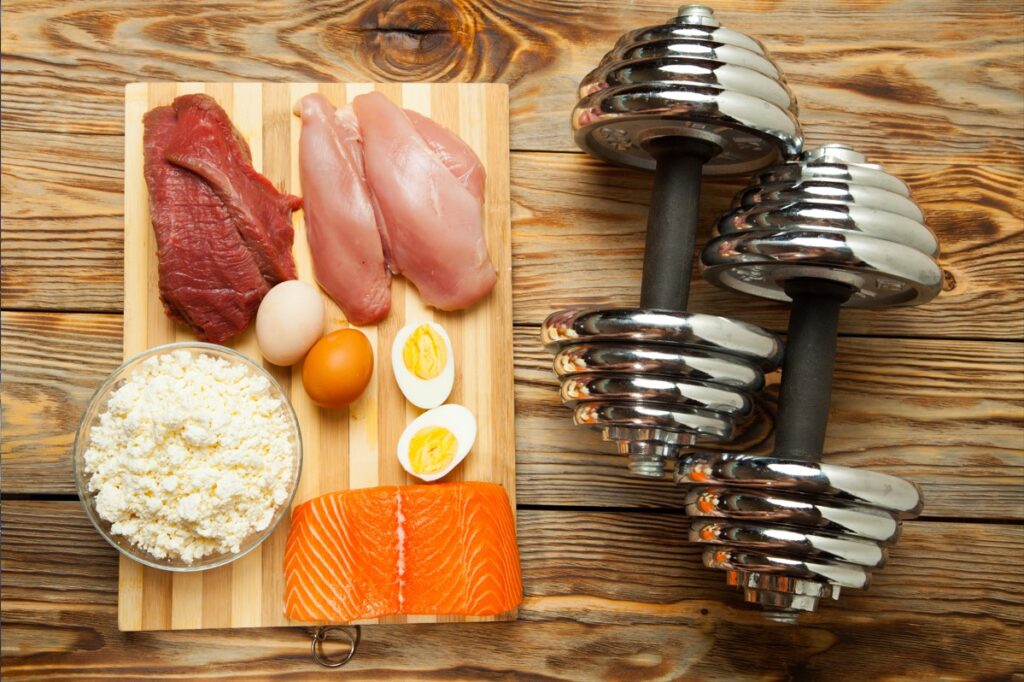 Platter of a healthy diet containing fish, cheese, eggs, meat, chicken and dumbbells on a wooden background.